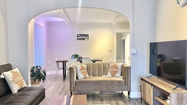 A modern living room with a sofa, coffee table, TV stand, and a dining area in the background.