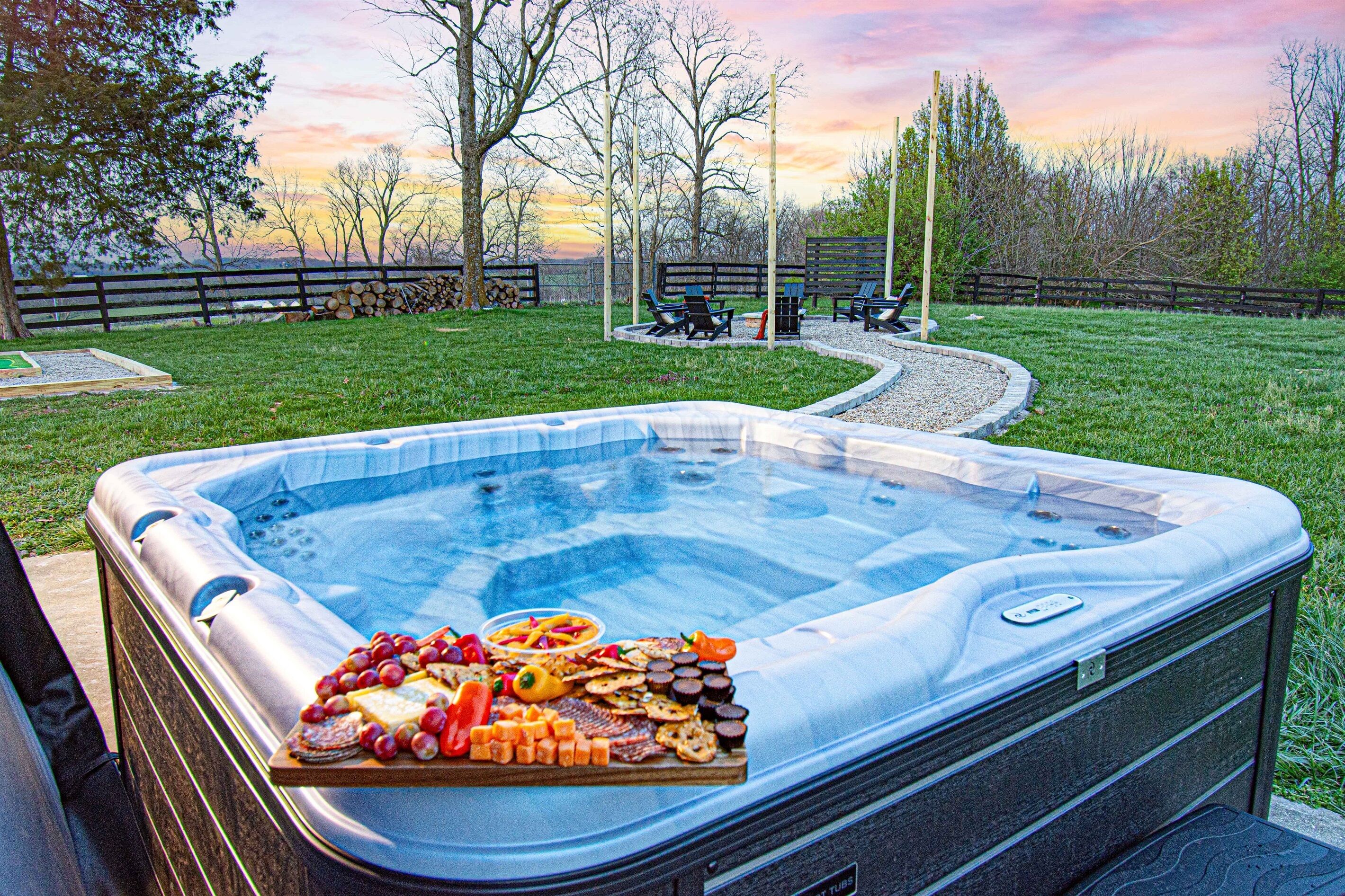 A hot tub with a variety of snacks on the edge, surrounded by a grassy area with benches and a wooden fence.