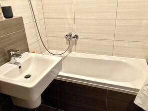 Une salle de bain avec un lavabo et une baignoire blancs, un flexible de douche et des murs recouverts de carreaux foncés.
