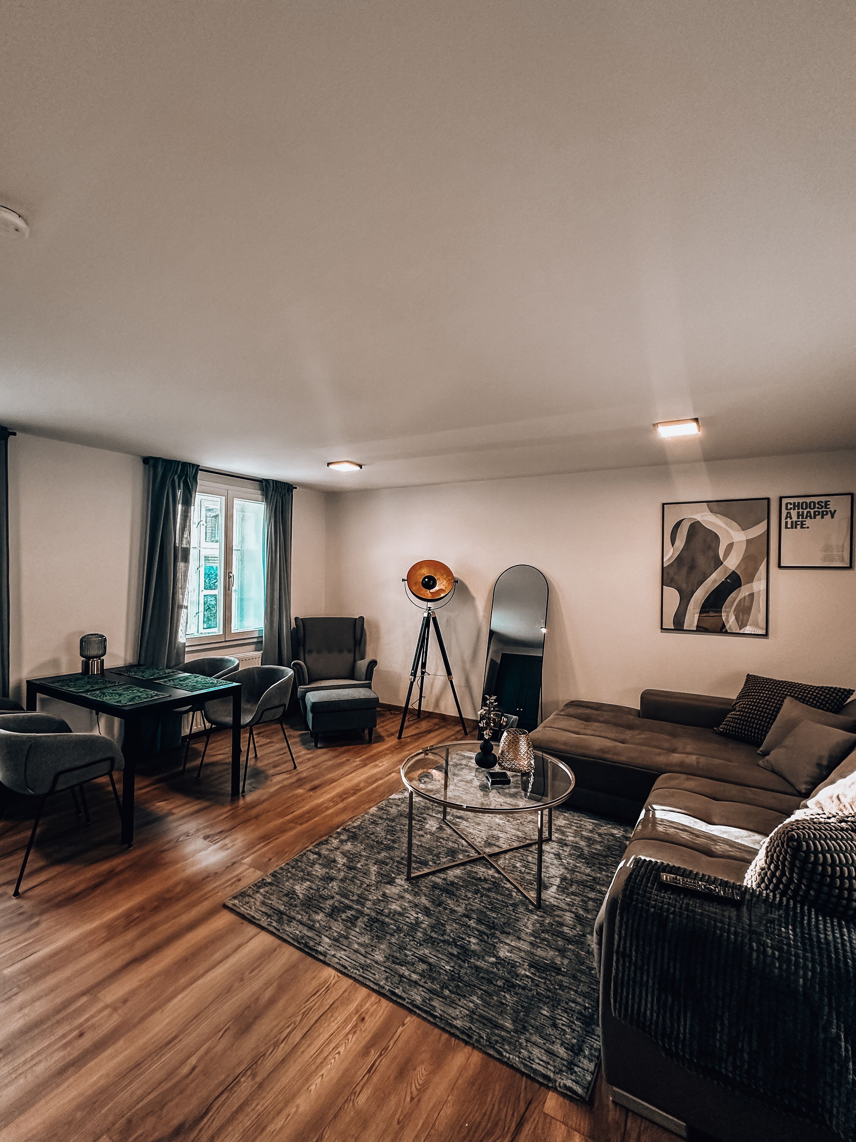 En moderne stue med trægulv, spiseplads med bord og stole, sofa, gulvlampe og to billeder i rammer på væggen.