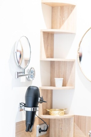 A bathroom with a hairdryer mounted on the wall, a mirror, and wooden shelves with a cup and a decorative bowl.