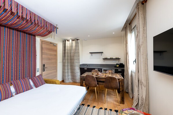 A bedroom with a bed, a dining table with chairs, a kitchen area, and a TV mounted on the wall.