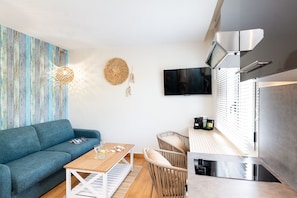 Un salon moderne avec un canapé bleu, une table basse en bois, des fauteuils en osier, un téléviseur et un coin cuisine doté d’une hotte en acier inoxydable.