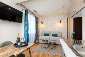 Un salon moderne comprenant un canapé, deux petites tables d’appoint, une table à manger en bois avec une bouteille et un verre, et un coin cuisine avec un évier.