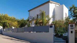 Un edificio blanco con techo plano y balcón, rodeado por un muro alto y vegetación.
