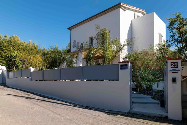 Un bâtiment blanc, doté d’un toit plat et d’un balcon, entouré d’un mur élevé et de verdure.