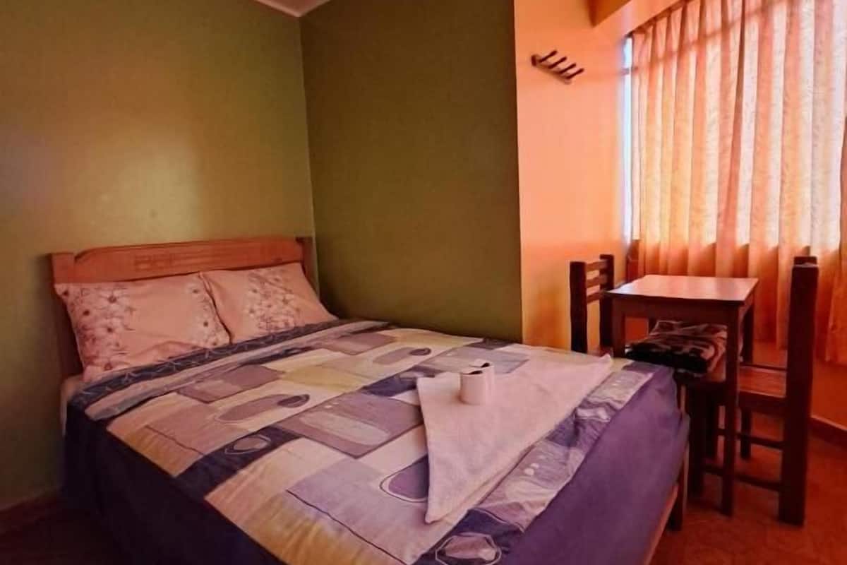 A single bed with a quilt, a small table, and a chair in a room with green walls and a window with curtains.