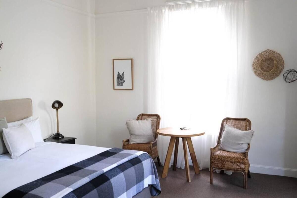 A hotel room with a bed, two wicker chairs, a small round table, a nightstand, a lamp, and a framed picture on the wall.