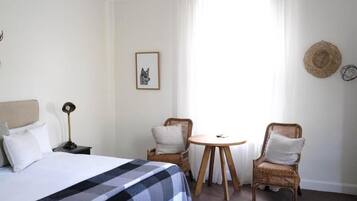 Habitación de hotel con cama, dos sillones de mimbre, una mesita redonda, una mesita de noche, una lámpara y un cuadro en la pared.