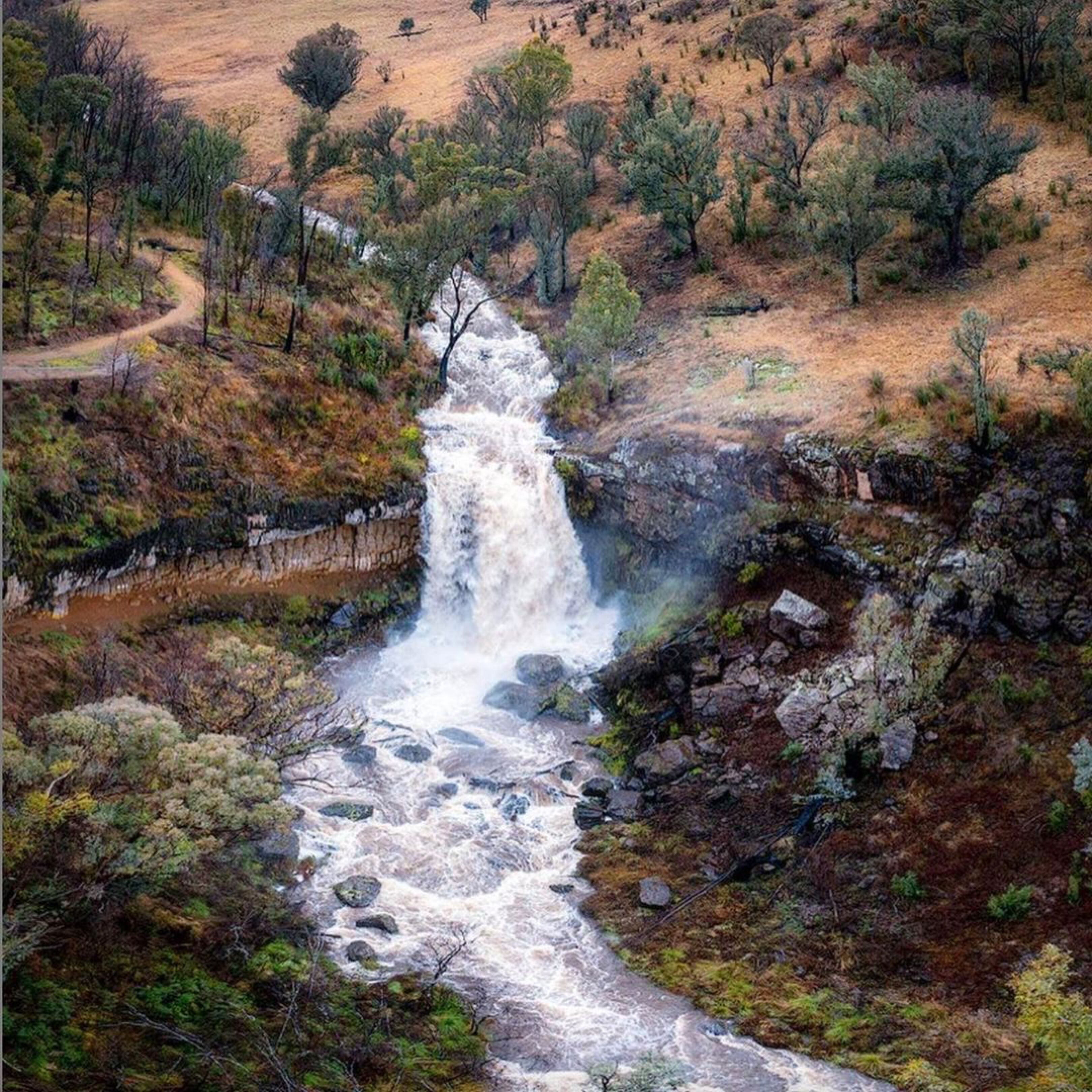 A waterfall cascading over rocks in a natural setting with surrounding trees and a dirt path.