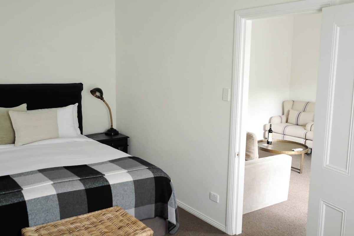 A bedroom with a bed, bedside table, lamp, and a chair in the adjacent living area.