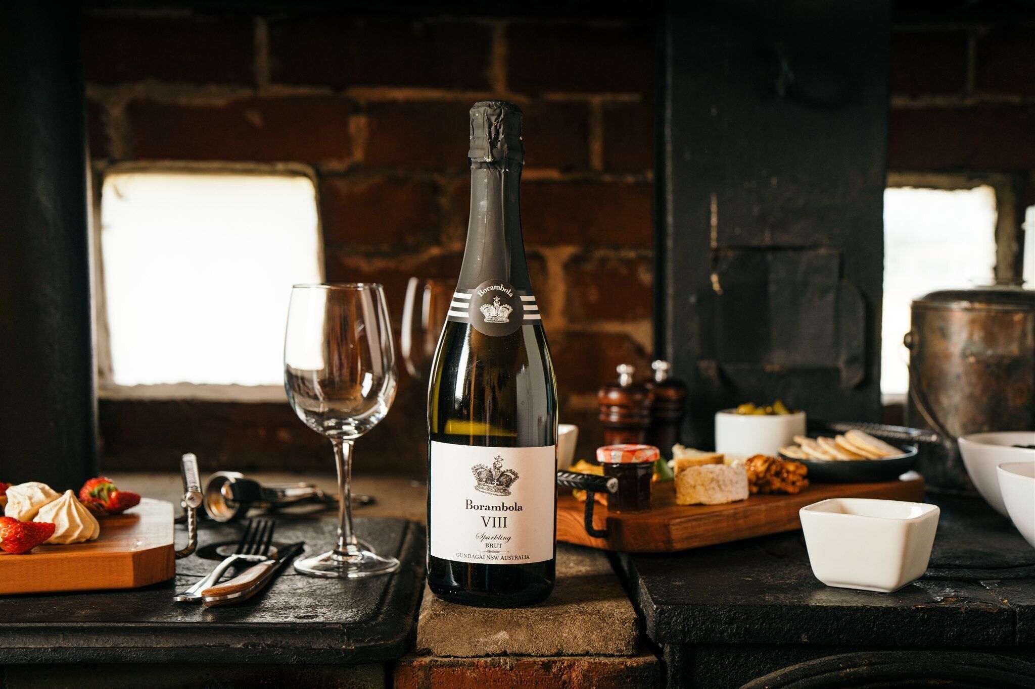 Une bouteille de vin mousseux Borambola VIII Brut est posée sur une table, entourée de verres, d’une planche en bois sur laquelle se trouvent des aliments, et un mur de briques rustiques en arrière-plan.