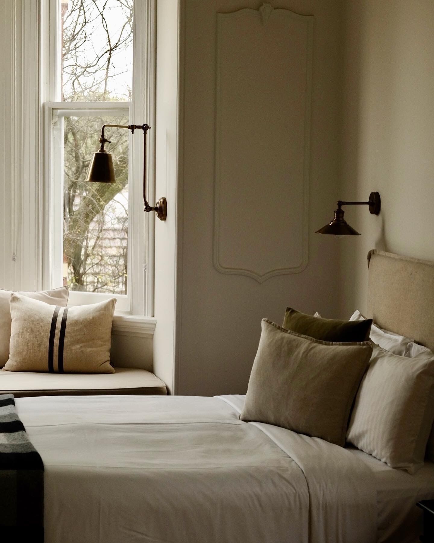 A bedroom with a large window, a bed with multiple pillows, a bedside table, and a wall-mounted lamp.