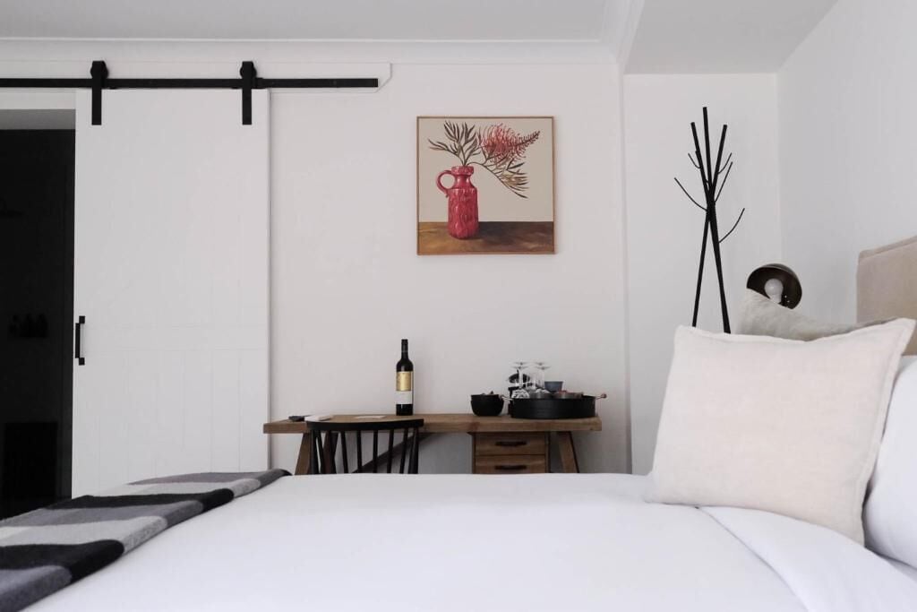 Un dormitorio con una cama, un escritorio con una botella y una tetera, un cuadro de un jarrón con flores, y un jarrón negro alto.