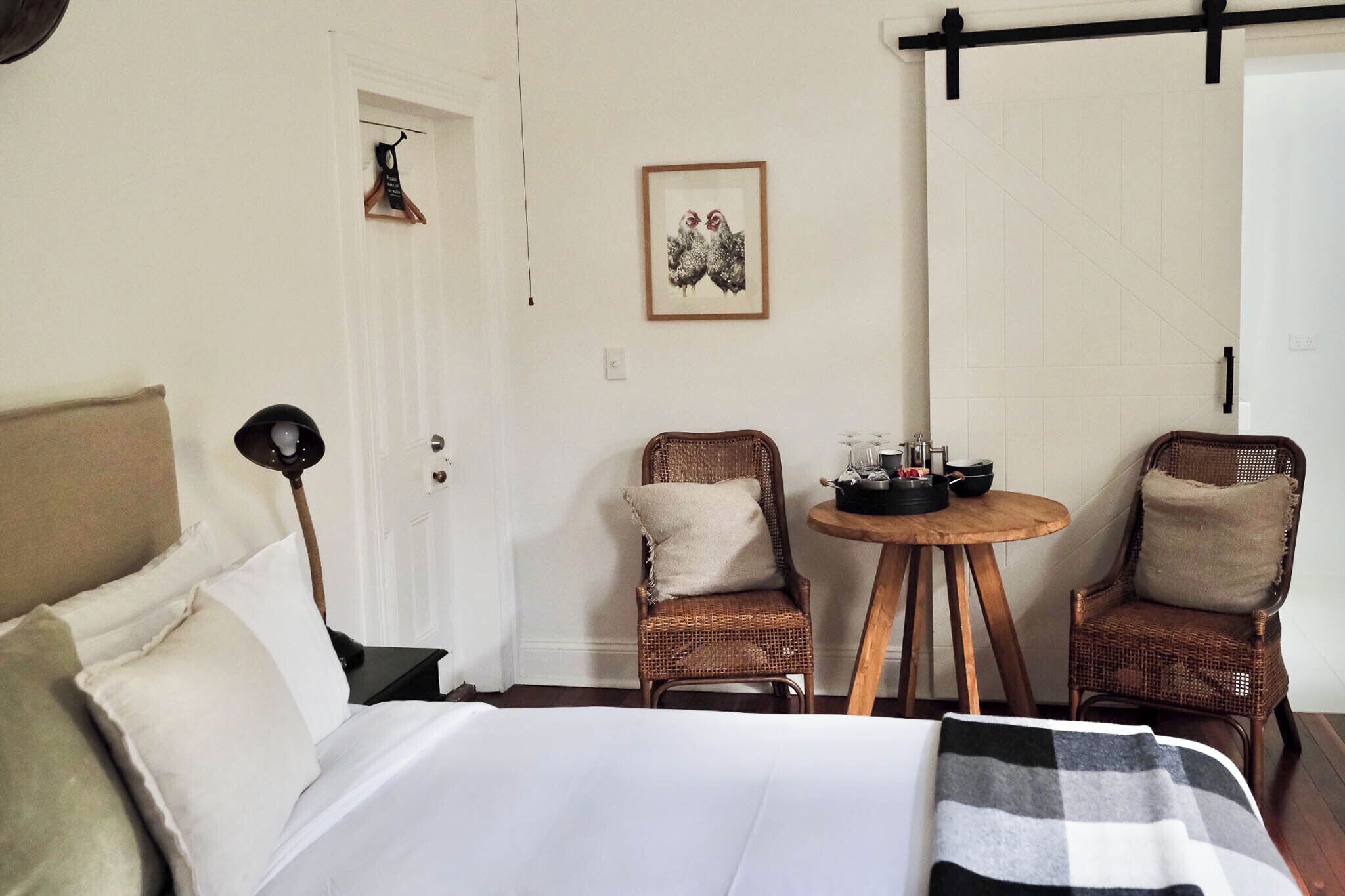 Quarto com cama, duas cadeiras de vime, uma mesinha redonda com um conjunto de chá e um quadro na parede.