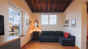 A room with a wooden floor, a dark sofa, a small table with a lamp, a TV, and framed pictures on the walls.