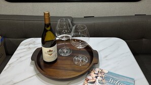 A table with a bottle of wine, two glasses, and a tray in a vehicle interior with a view of greenery through the window. - Beach Haven (Sekiu)
