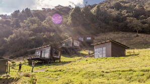 A hillside with modern, elevated wooden structures, possibly cabins or houses, with a clear sky and sunlight. - Fuga Neusa (Tausa)