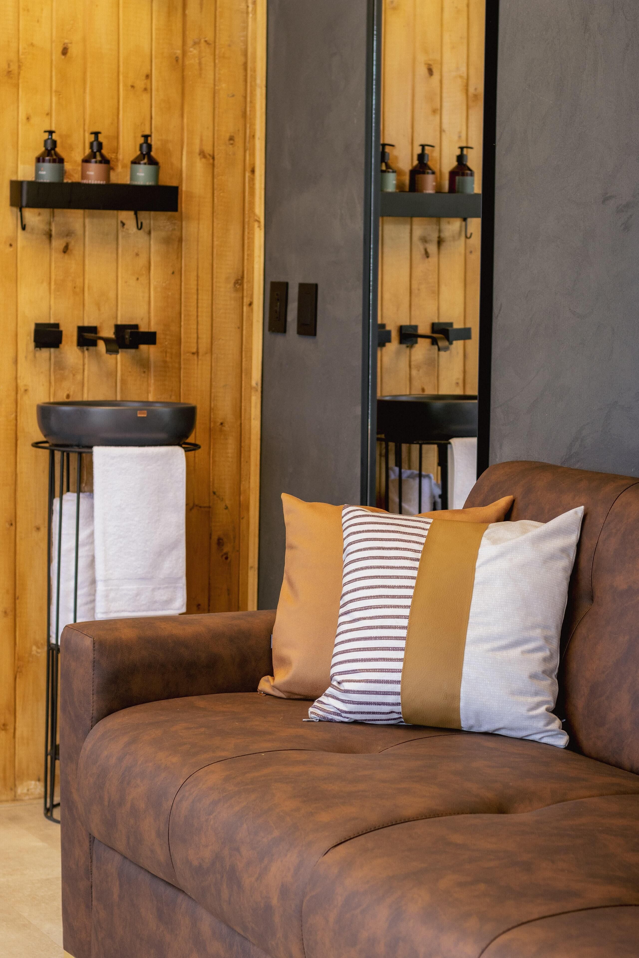 A brown leather sofa with striped and solid pillows, a wooden wall with shelves holding toiletries, and a towel rack with a sink.