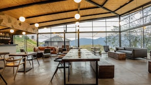 A spacious indoor area with a long wooden table, multiple chairs, and a sofa. Large windows provide a view of the outdoors. - Fuga Neusa (Tausa)