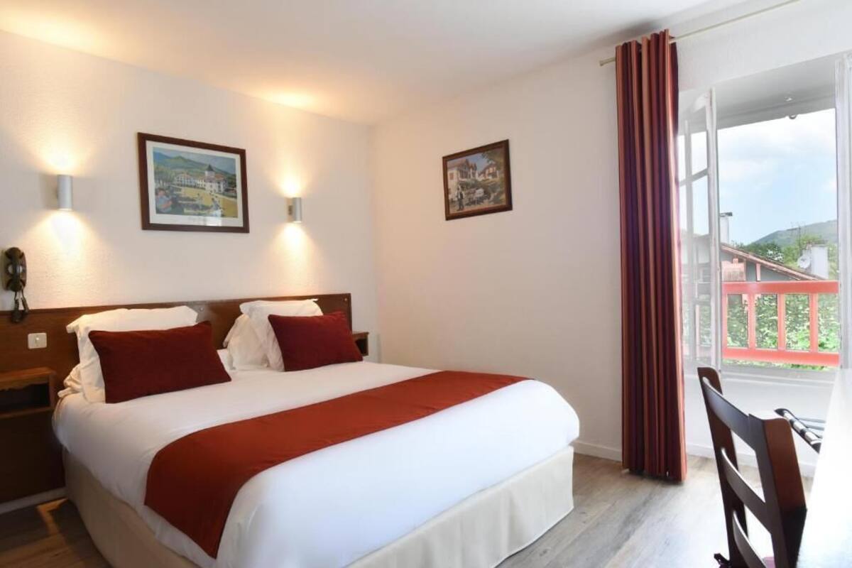 A hotel room with a bed, two wall-mounted lights, two framed pictures, a window with curtains, and a view of a balcony.