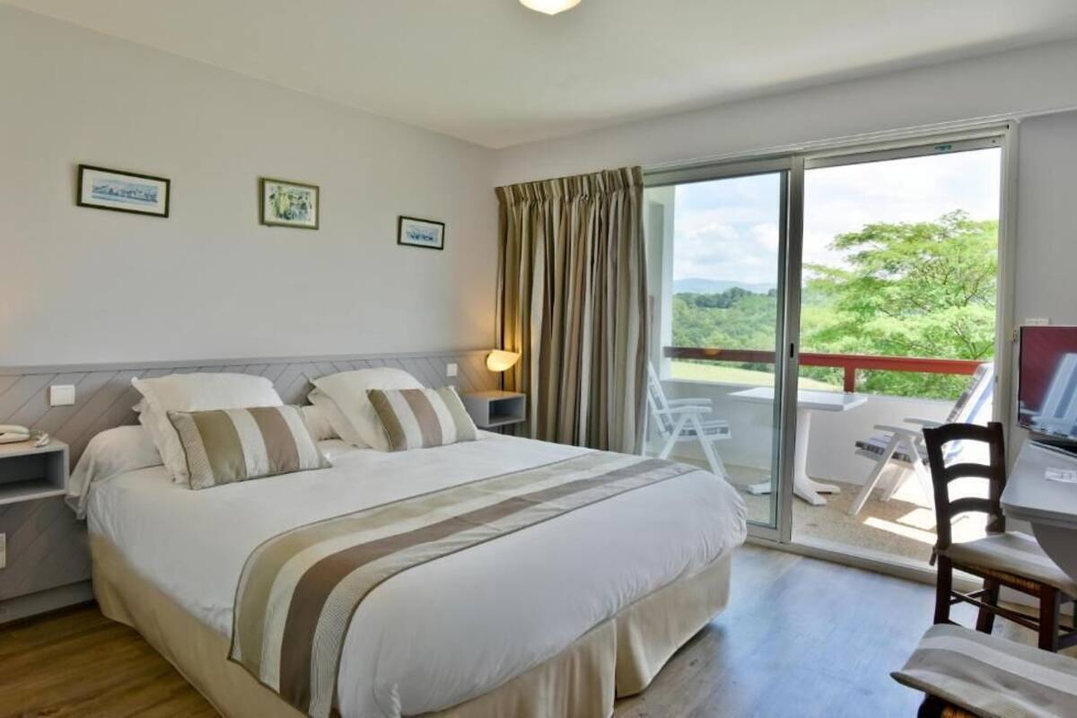 A bedroom with a bed, bedside tables, a desk, and a chair. There is a balcony with a view of greenery.