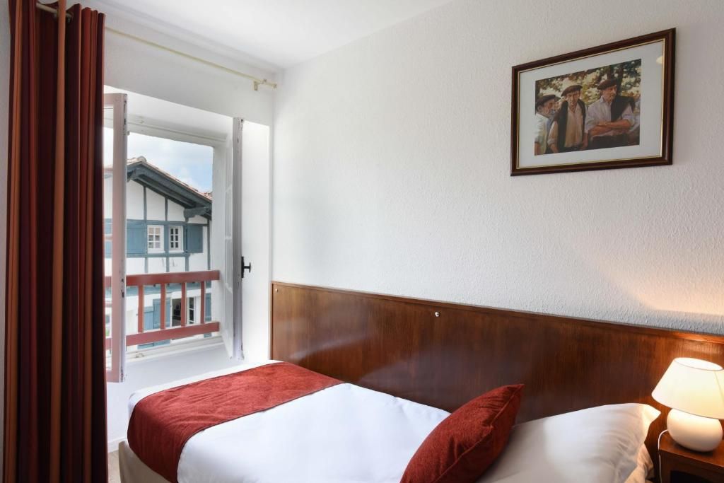 Une chambre d’hôtel avec un lit, une table de chevet avec une lampe, un tableau encadré au mur et une vue sur un bâtiment à l’extérieur.