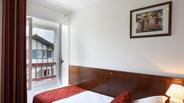 A hotel room with a bed, a nightstand with a lamp, a framed picture on the wall, and a view of a building outside. - Logis Hotel Baratxartea (Sare)