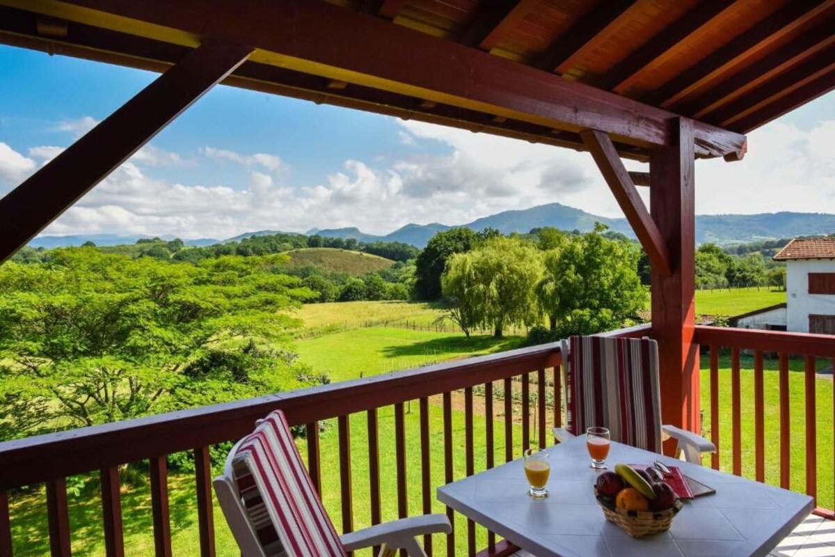 A balcony with a table, chairs, and a view of green fields and mountains.