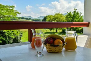 A table with a glass pitcher of orange juice, a glass of pink drink, and a basket of fruits on a balcony with a view of green fields and mountains. - Logis Hotel Baratxartea (Sare)