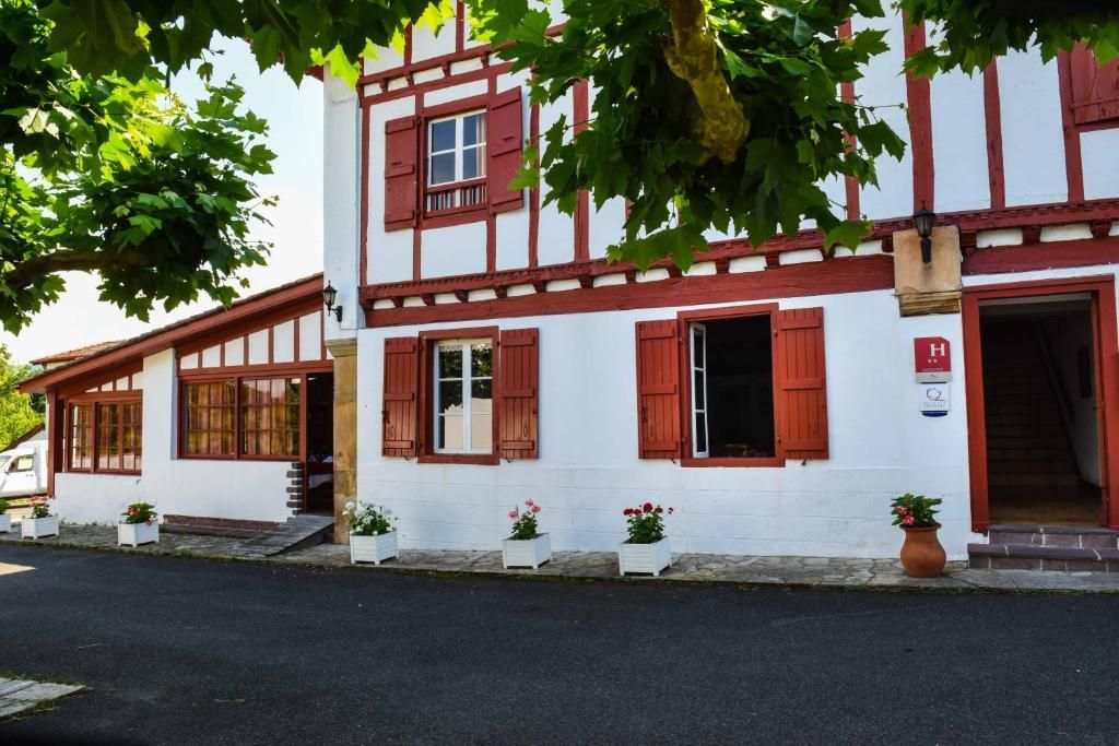 Un edificio tradicional de entramado de madera con contraventanas rojas, paredes blancas y un pequeño porche con plantas en macetas.