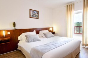 A neatly made bed with white linens and two pillows, a wooden headboard, bedside tables with lamps, and a painting on the wall. - Logis Hotel Baratxartea (Sare)