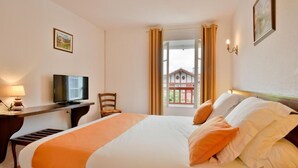 A hotel room with a bed, a desk with a television, a chair, and a window with curtains. - Logis Hotel Baratxartea (Sare)