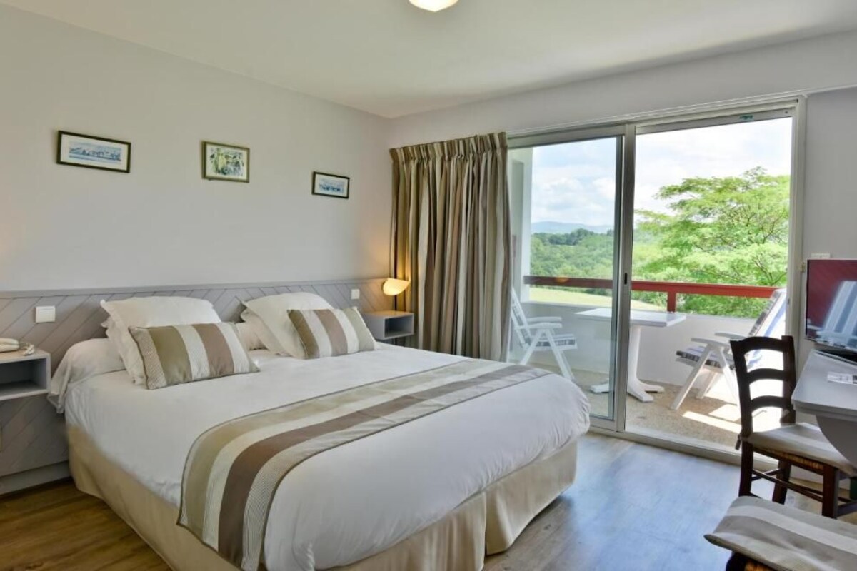 A bedroom with a bed, bedside tables, a desk, and a chair. There is a balcony with a view of greenery.