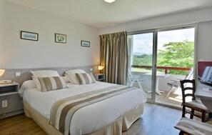 A bedroom with a bed, bedside tables, a desk, and a chair. There is a balcony with a view of greenery. - Logis Hotel Baratxartea (Sare)
