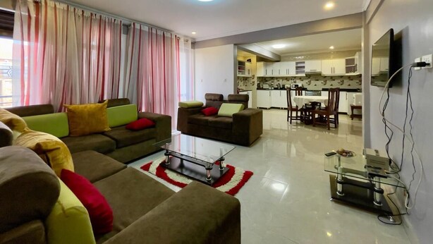 A modern living room with a sofa set, a glass coffee table, and a dining area with a kitchen in the background. - Executive 2 Bed Apartment in Kigali Kagarama (Kigali)