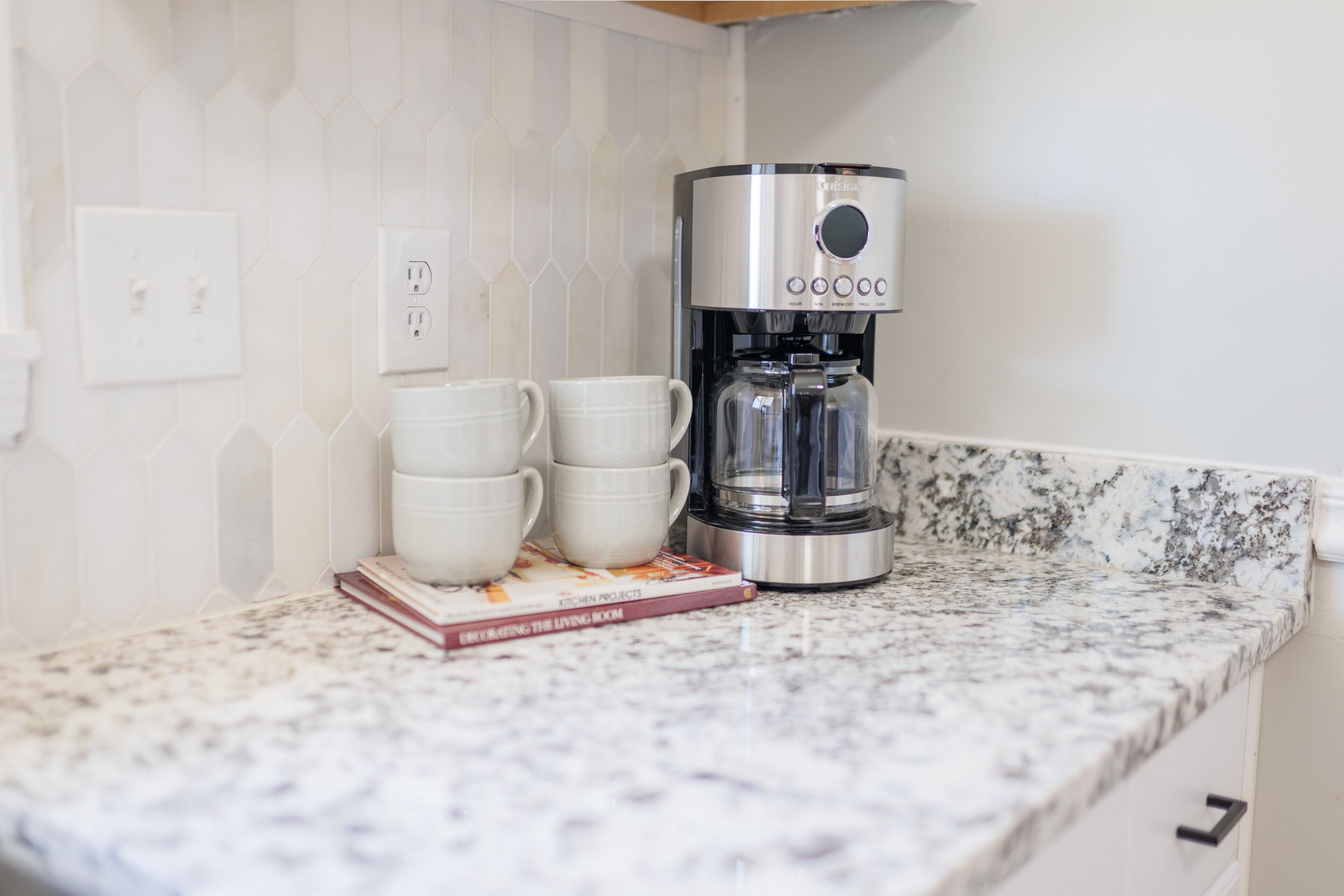 Une cuisine équipée d’une machine à café, de trois tasses blanches et d’un livre de recettes posé sur le comptoir.