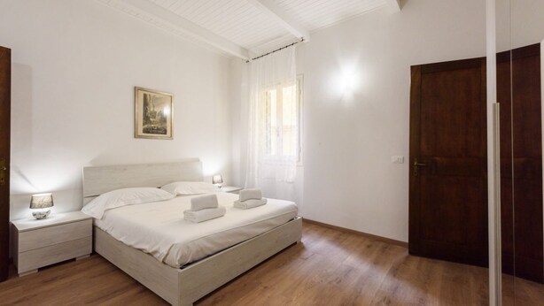 A bedroom with a bed, nightstands, a wooden floor, a door, and a window with curtains. - Uffizi 3 bedrooms in Florence by Mmega (Firenze)