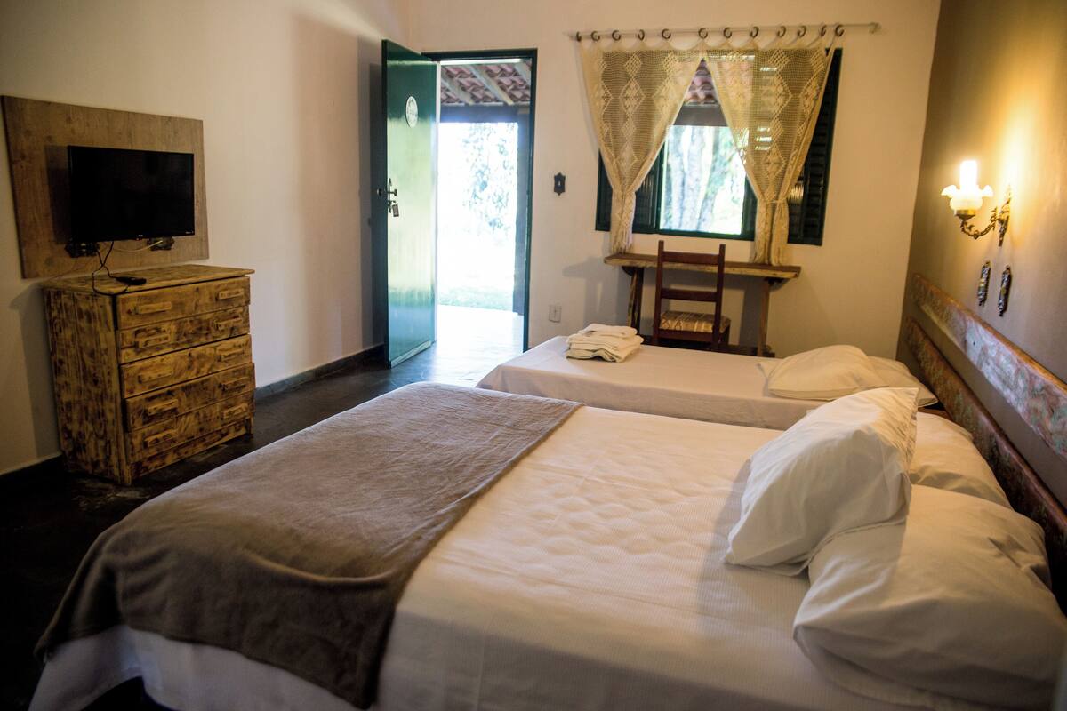 A hotel room with two beds, a TV on a wooden dresser, a window with curtains, and a wall-mounted light fixture.