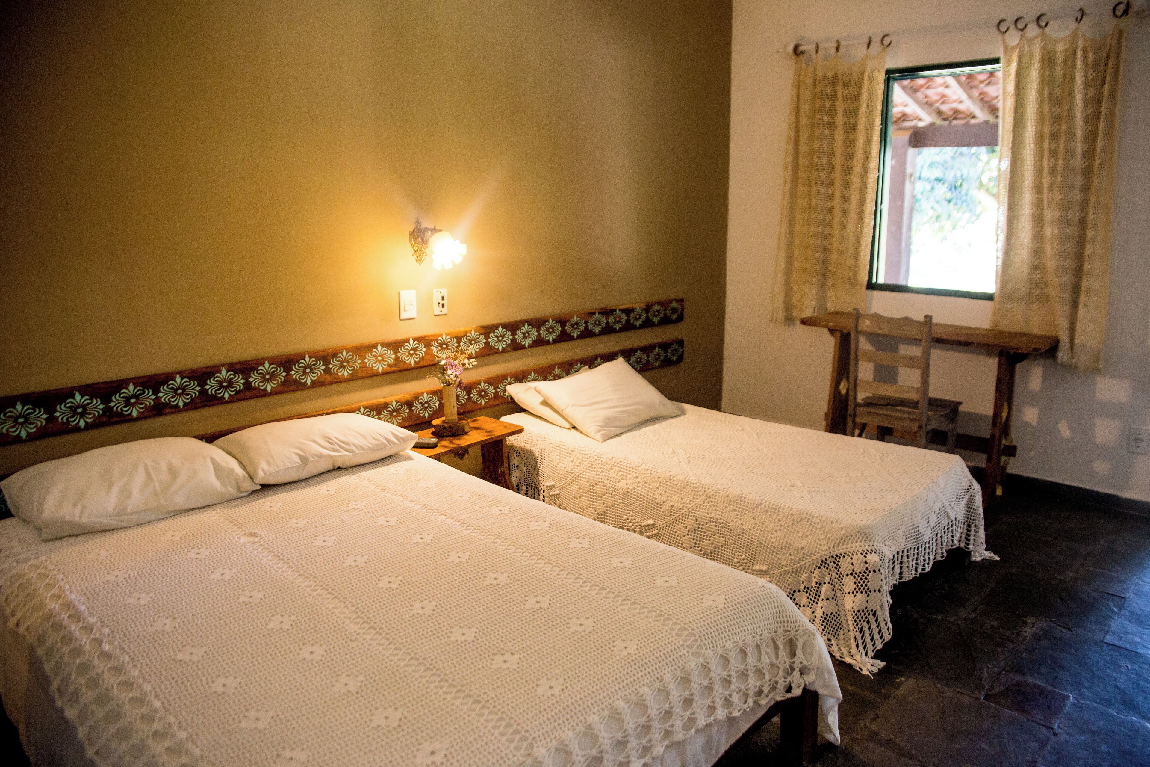 A room with two beds, a wooden headboard with floral patterns, a nightstand with a lamp, and a window with curtains.