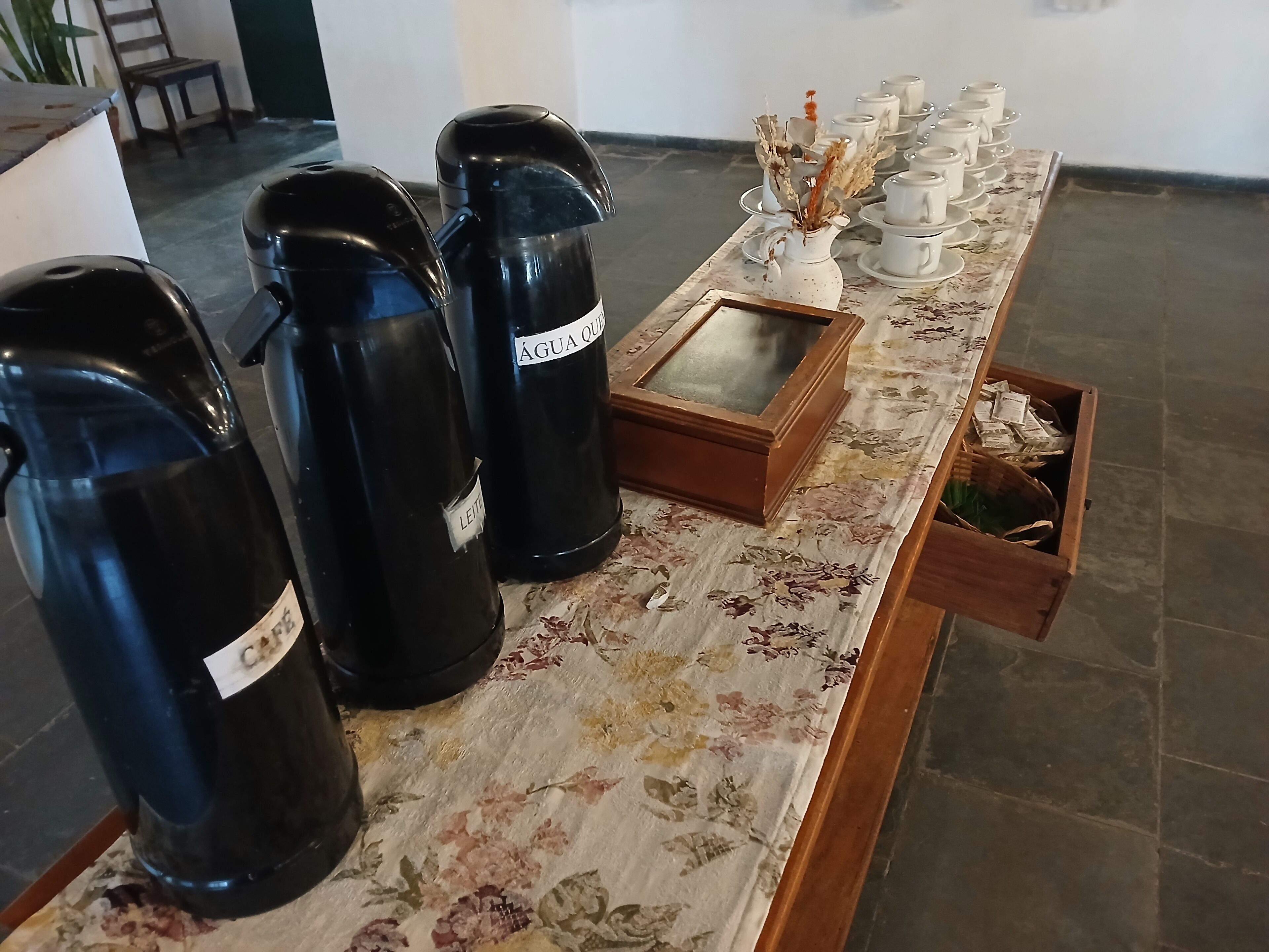 A table with black containers labeled "Café" and "Leite," a white pitcher with dried flowers, and a tray with white cups and saucers.