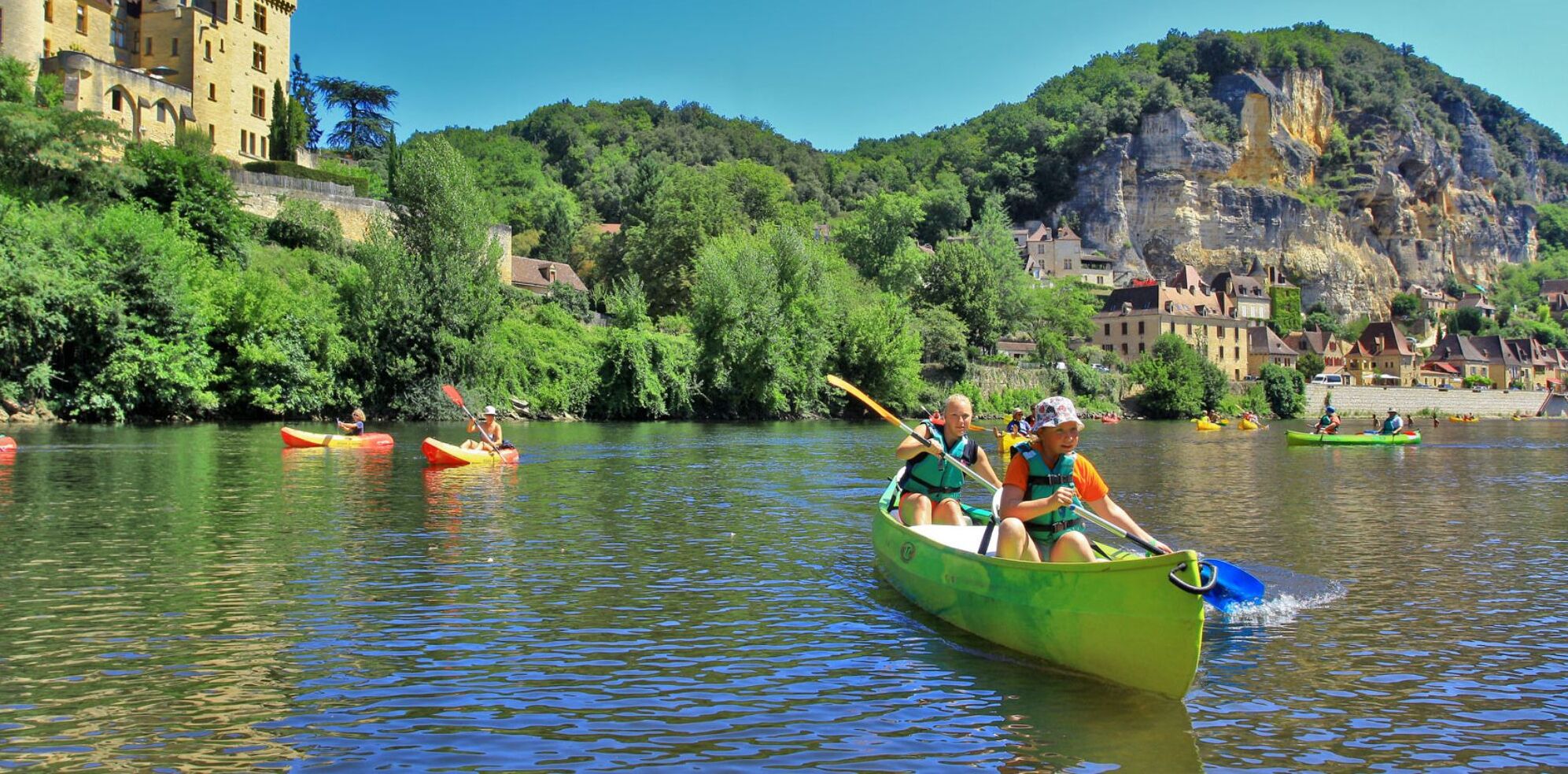 A river with people kayaking. There are buildings and trees along the riverbank.