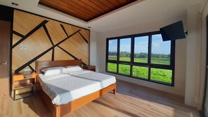 A modern bedroom with a large bed, wooden flooring, a sloped ceiling, and a large window offering a view of a green field. - CWC Solo/Duo Base: King Suite & Shared Luxury | Walk to CWC (Pili)