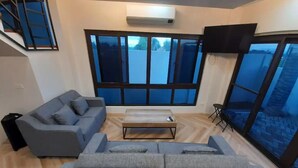 Ein modernes Wohnzimmer mit einer grauen Couch, einem Couchtisch und einem großen Fenster mit Blick auf blauen Himmel und Bäume.