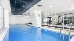 Ein moderner Innenpool mit großen Glaswänden, blauen Wasseroberflächen und einem durch die Scheiben einsehbaren Fitnessbereich.