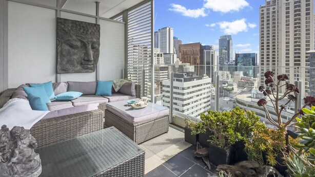 Ein moderner Balkon mit Korbmöbeln, einer Steinskulptur und Blick auf die Stadt.