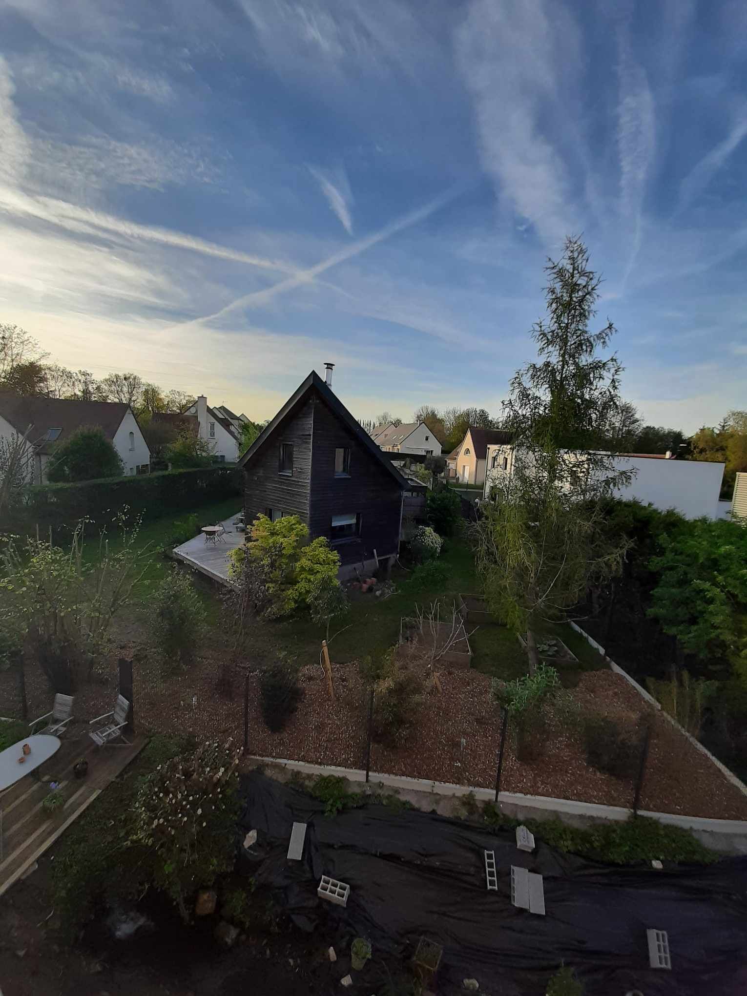 Un barrio residencial con una casa oscura de techo inclinado y chimenea, rodeado de follaje y un pequeño estanque.