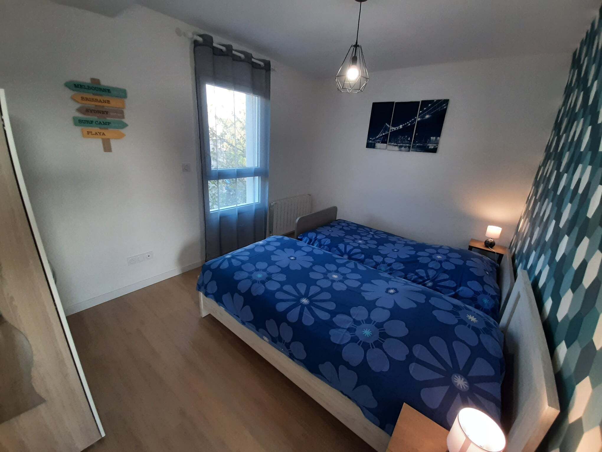 A bedroom with a bed, a window with curtains, a wall-mounted sign with city names, and a framed picture.