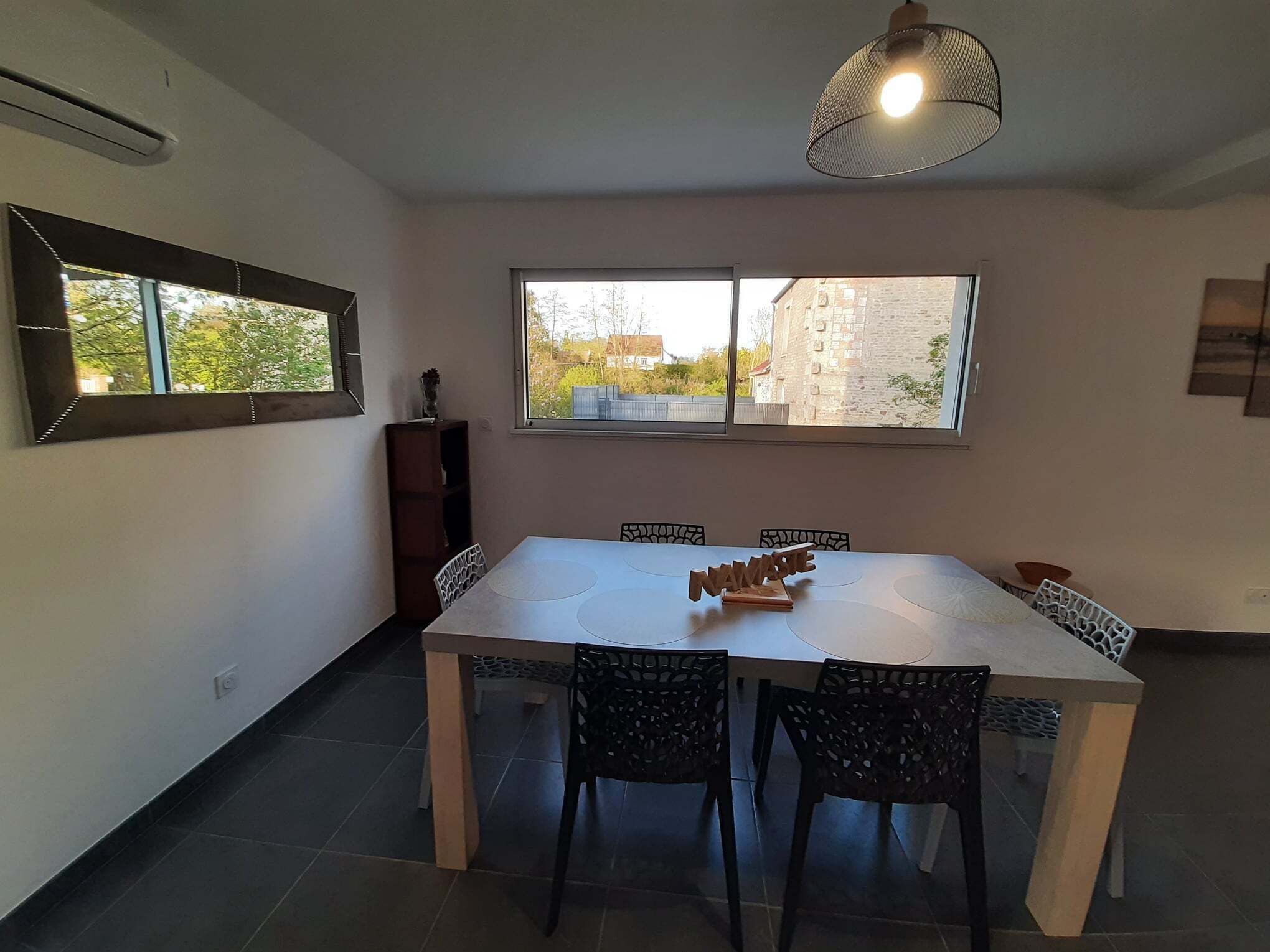 A dining area with a white table and black chairs, a large window with a view of buildings, and a hanging light fixture.