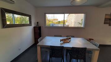 A dining area with a white table and black chairs, a large window with a view of buildings, and a hanging light fixture.